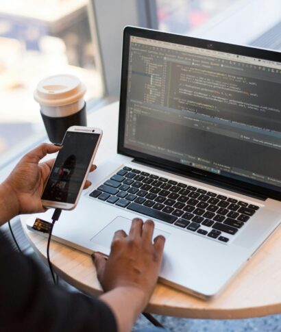 pexels-photo-1181244-1181244 Developer working remotely, coding on a laptop with phone in hand, showcasing modern work culture.