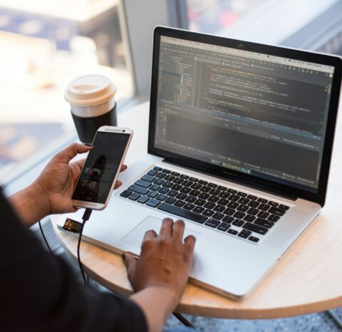 Developer working remotely, coding on a laptop with phone in hand, showcasing modern work culture.
